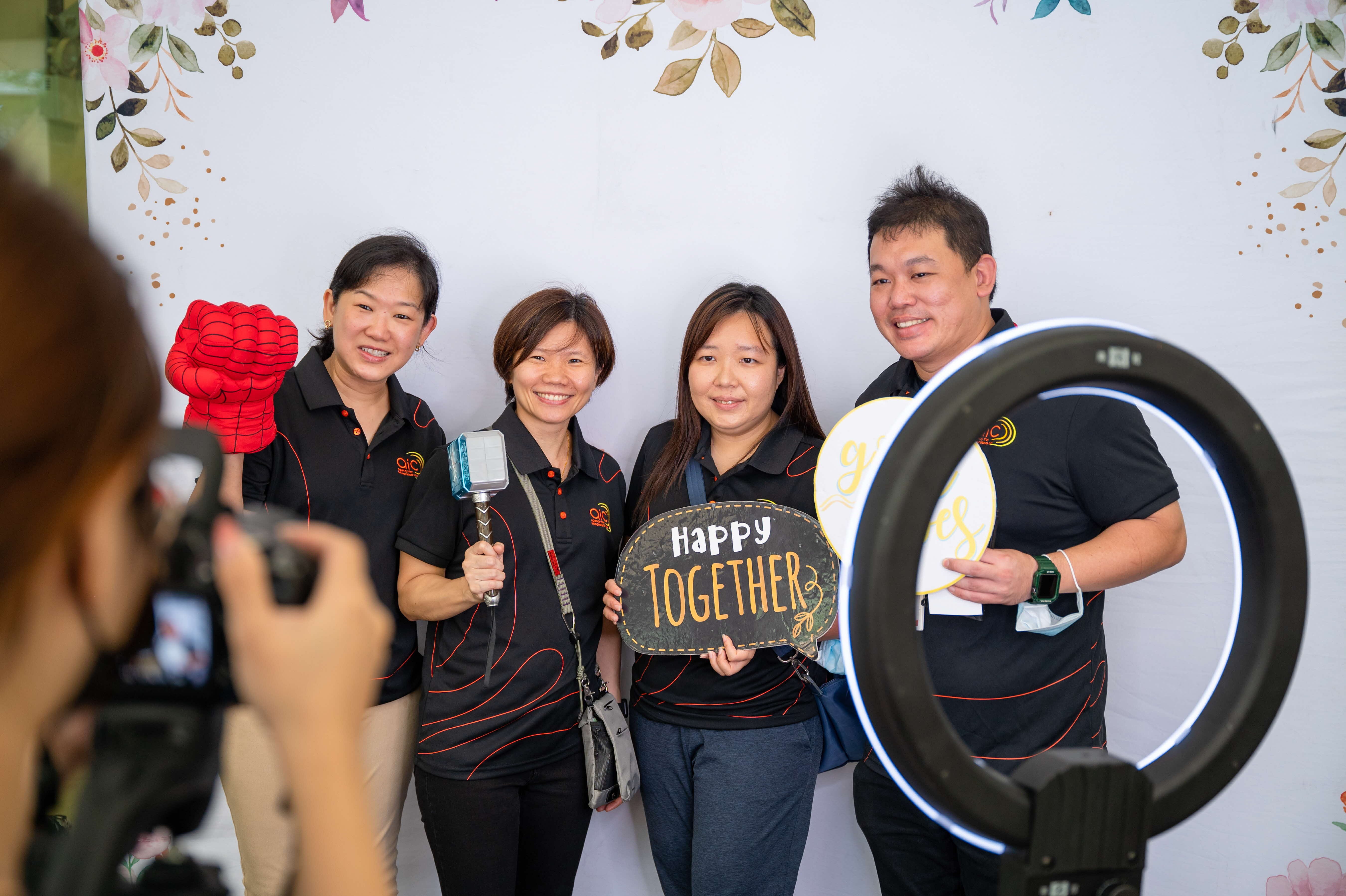 Four people in matching black uniforms pose with props, signs, and a ring light; one holds a "Happy Together" sign.